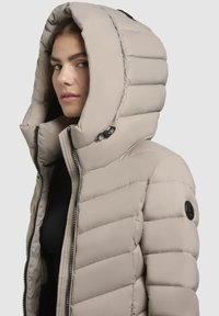 Bežna puhovka z velikim kapuco, navpično prešita tekstura, zadrga na sprednji strani in logotipom na rokavu.