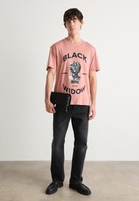 Světle růžové tričko s černým grafickým potiskem s nápisem "BLACK WIDOW EST 1993." Černé džíny, černá taška a černé boty doplňují outfitu.