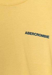 Kollane puuvillane t-särk, millel on ümmargune kaelus ja esiküljel trükitud sinine "ABERCROMBIE" logo. Sujuv tekstuur, casual disain.