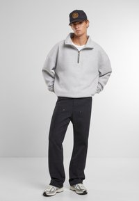 Pulóver de felpa gris con cuello con cremallera, combinado con pantalones oscuros con acentos dorados y completado con zapatillas multicolores y una gorra negra.