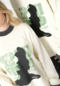 Kremowy, oversize'owy sweter z zielonym wzorem graficznym i czarnym nadrukiem sylwetki. Czarny ściągacz przy mankietach i kołnierzu.