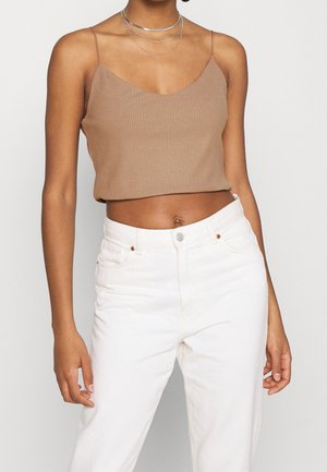 Person trägt ein ärmelloses, beiges, geripptes Crop-Top, weiße, hoch taillierte Hosen und geschichtete silberne Halsketten vor einem einfarbigen Hintergrund.