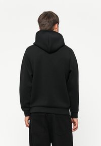 Un homme portant un sweat à capuche noir uni et un pantalon noir, vu de dos contre un fond blanc uni.