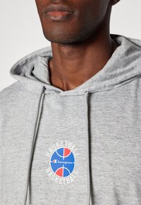 Pelēks kokvilnas kapucis ar priekšējo logotipu, kas attēlo sarkanu, zilā un baltā krāsā veidotu basketbola dizainu. Iekļauti saišķi un teksturēta auduma apdare.