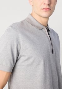 Šedá polo tričko s krátkymi rukávmi, zipsovým výstrihom a kontrastným golierom. Hladká textúra s minimálnymi švovými detailmi. Jednoduchý a klasický dizajn.