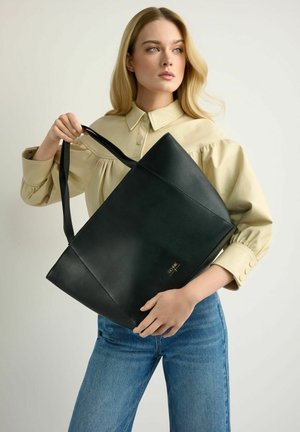 Kobieta w beżowej bluzce i niebieskich dżinsach trzyma dużą czarną skórzaną torebkę na ramię, spoglądając w bok na jasnym tle.