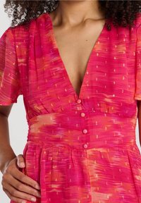 Roze jurk met een diepe V-hals, korte mouwen en een gestructureerde stof met een abstract patroon. Knopen aan de voorkant voegen detail toe aan het ontwerp.