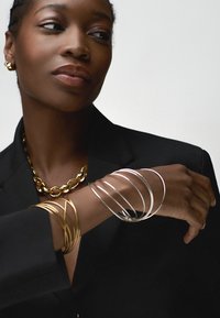 Collier en or, plusieurs bracelets fins en argent et en or sur une peau foncée. Porté avec un blazer noir. Accent sur les détails et textures des bijoux.