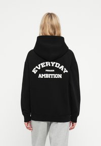 Čierna mikina s kapucňou, na zadnej strane s veľkým bielym nápisom v zakrivenom písme "EVERYDAY AMBITION". Štandardný strih s kapucňou.
