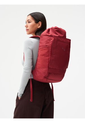 Frau in hellgrauem Langarmshirt und dunkler Hose mit großem rotem Rucksack, die seitlich vor weißem Hintergrund steht.