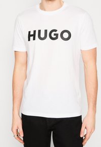 Biała bawełniana koszulka z krótkim rękawem z odważnym czarnym nadrukiem "HUGO" na piersi. Standardowy krój z okrągłym dekoltem.