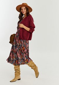 Długa kwiatowa sukienka na ciemnym tle, kolorowe kwiaty; bordowy, oversize'owy sweter; beżowe, luźne kozaki za kolano; brązowa torba; kapelusz z szerokim rondem.