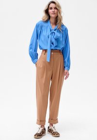 Blauwe blouse met strikdetail aan de voorkant, losse fit en geplooid schouderepauletten. Bruine plooibroek met hoge taille en sluiting met twee knopen. Sneakers met luipaardprint.