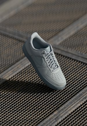 Gray Nike sneaker floating above textured metal grid surface, showing side and top views with visible laces and perforations on toe box.