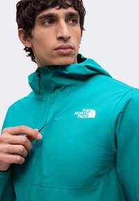 Jeune homme ajustant le cordon de la veste à capuche turquoise North Face sur un fond uni.