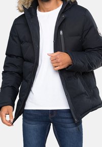 Sort dunjakke med pelsforet hætte, lynlås og knaplukning samt forlommer, båret over en hvid skjorte og mørke jeans.