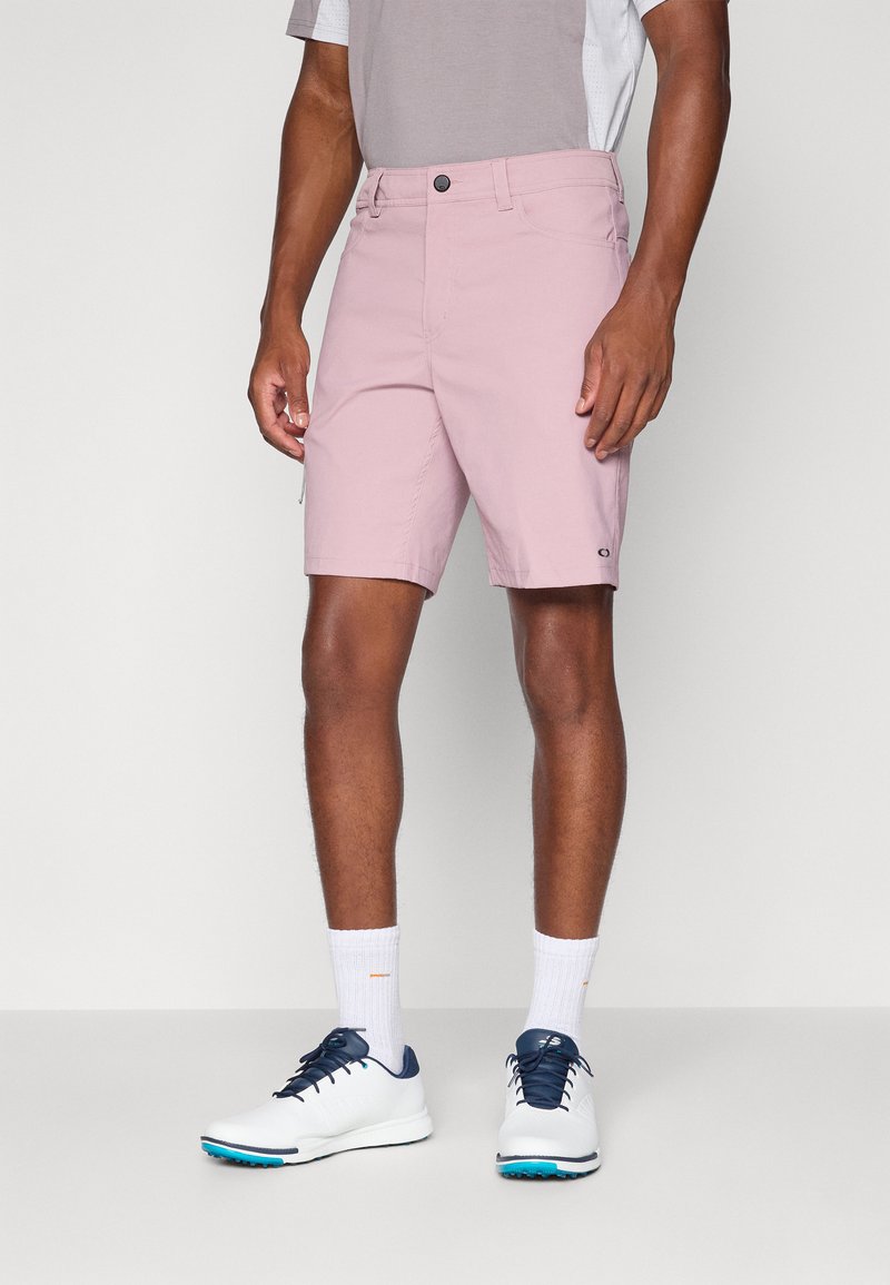 Light pink shorts with a button closure, front pockets, and subtle logo detail. Paired with white shoes and ribbed socks.