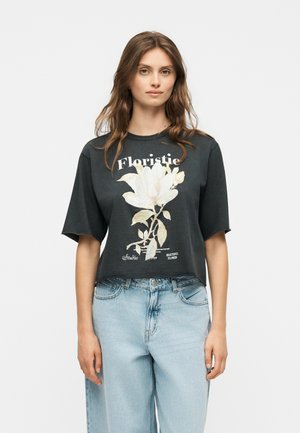 Jeune femme portant un t-shirt court noir avec un grand motif de fleur blanche et du texte, associé à un jean taille haute bleu clair.
