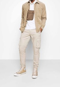 Beige corduroy overhemd over een witte polo, gecombineerd met lichtbeige cargobroek en lichtbruine hoge sneakers. Een bruine pouch hangt aan het overhemd.
