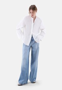 Camicia bianca con colletto alla mandarino, abbinata a jeans a gamba larga di colore azzurro chiaro. Tessuto morbido e vestibilità rilassata.