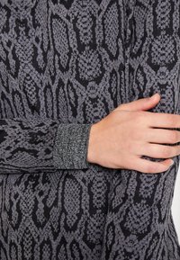 Tessuto con motivo a serpente nero e grigio con polsino testurizzato. Primo piano che mostra i dettagli del design e le mani sovrapposte sulla manica.