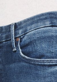 Jeans de mezclilla azul oscuro con efecto desgastado, que muestran un primer plano de la cinturilla, los pasadores del cinturón y un bolsillo delantero.