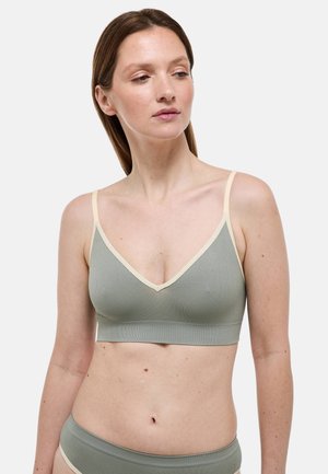 Femme à la peau claire et aux cheveux bruns portant un soutien-gorge côtelé vert sauge et une culotte assortie avec une bordure crème, debout devant un fond blanc uni.