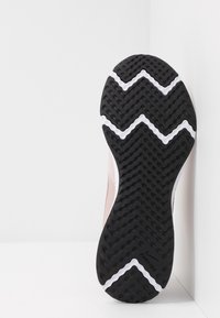 Semelle d'une chaussure de course avec une bande de roulement texturée noire et des lignes blanches en zigzag, appuyée contre un mur blanc en angle.