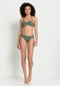 Woman wearing a green two-piece swimsuit with halter top and side-strap bikini bottoms, standing barefoot against a plain white background.