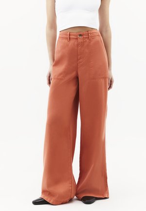 Mujer que lleva pantalones de tiro alto, pierna ancha en naranja óxido con bolsillos parche delanteros, combinados con zapatos negros puntiagudos y un top corto blanco.
