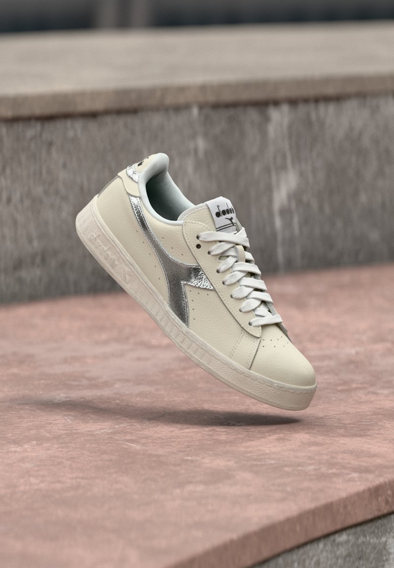 Λευκό αθλητικό παπούτσι με ασημί πλαϊνή ρίγα και κορδόνια, αιωρούμενο πάνω από μια πέτρινη επιφάνεια με φόντο έναν θολό γκρι τοίχο.