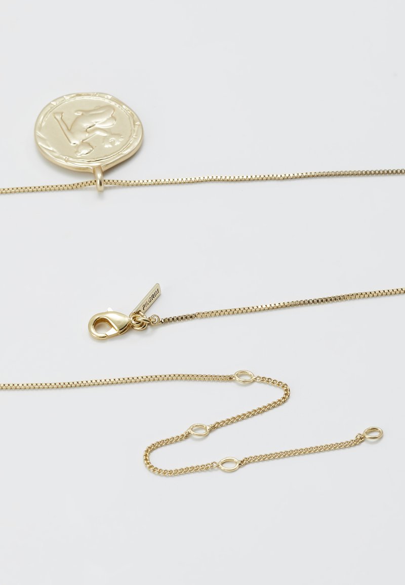 El collar en tono dorado presenta un colgante de moneda redonda con grabado, una cadena de eslabones y un cierre de langosta. Destaca por su textura suave y su detalle.