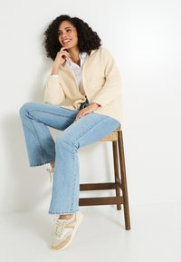 Cardigan beige avec une ouverture devant, assorti à un jean évasé bleu clair et des baskets beiges. Mannequin assis sur un tabouret en bois contre un fond clair.
