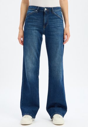 Straight-leg blue jeans worn by a person standing, paired with white sneakers and a white top, shown from mid-torso to feet.