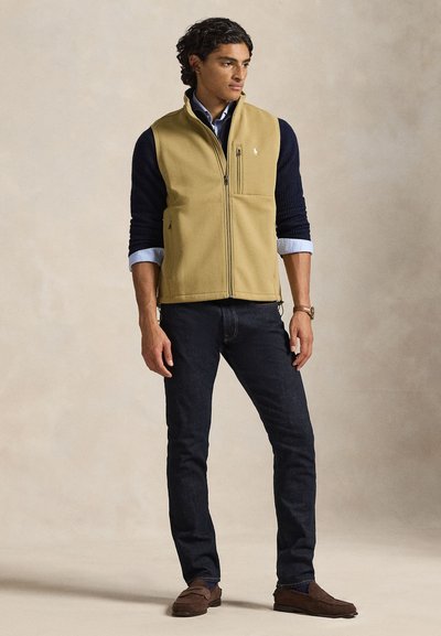 Beige fleece-liivi, jossa täysipitkä vetoketju ja pystykaulus, sekä sen alla sininen pitkähihainen paita. Tummat farkut ja ruskeat loaferit täydentävät asun.