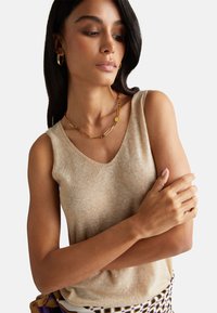 Beige bezramenný pletený top s výstřihem do V, jemnou texturou a přiléhavým střihem. Nosený se zlatými šperky, vyzařující čistý, minimalistický vzhled.