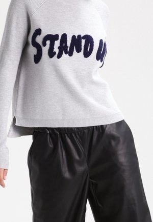 Szary sweter z napisem "STAND UP" dużymi czarnymi literami, zestawiony z luźnymi czarnymi skórzanymi spodniami na osobie na jednolitym tle.