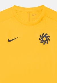 Gul atletisk skjorte laget av mykt, pustende stoff. Har en mørk logo og et sirkulært emblem på brystet. Rund halslinning.