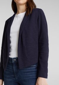 Blazer bleu marine, texturé, avec un col châle, ouverture sur le devant et deux poches frontales, porté sur une chemise blanche et un jean sombre.