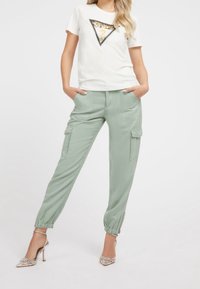 Camiseta blanca con un diseño triangular de lentejuelas, combinada con pantalones cargo verde claro con puños elásticos y bolsillos laterales. Tacones altos plateados.