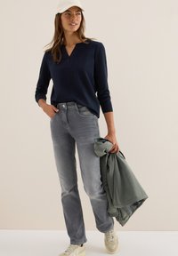 Kvinna klädd i marinblå långärmad topp, grå jeans, vita sneakers och vit keps, som håller en vikt grön jacka i sin högra hand.