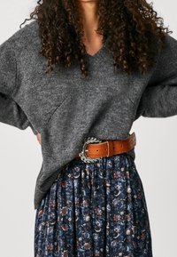 Grauer gestrickter Pullover mit V-Ausschnitt und überschnittenen Schultern, kombiniert mit einem blauen, floralen, plissierten Rock, akzentuiert durch einen braunen Ledergürtel mit einer gemusterten Schnalle.