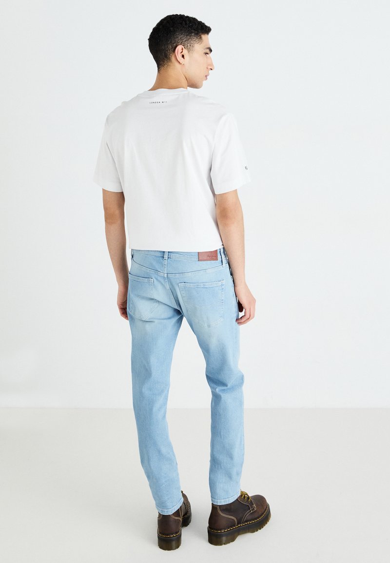 Weißes Baumwoll-T-Shirt mit kurzen Ärmeln und dem Text "LONDON WIN" auf dem Rücken, kombiniert mit hellblauen Jeans und braunen Schnürstiefeln.