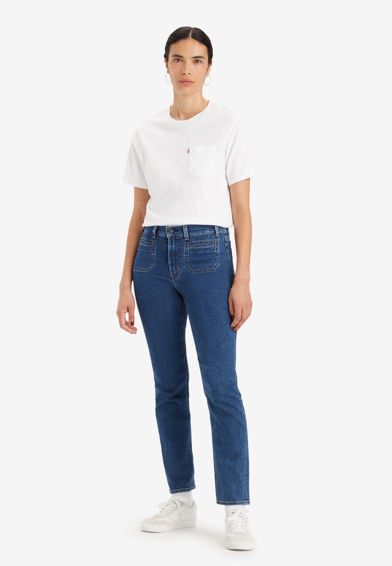 T-shirt en coton blanc avec une poche poitrine, associé à un jean en denim bleu taille haute et coupe slim, porté avec des baskets blanches.