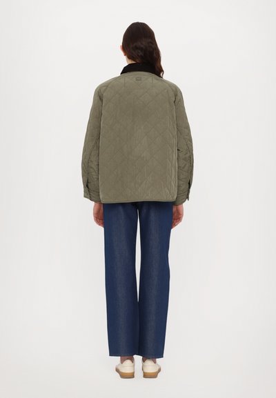 Olivgrüne gesteppte Jacke mit schwarzem Kragen, die ein Rautenmuster und eine lockere Passform aufweist, kombiniert mit weit geschnittenen blauen Denim-Hosen und weißen Schuhen.