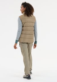 Beige Steppweste mit hohem Kragen und gestepptem Design, getragen über einem hellblauen Langarmshirt und kombiniert mit hellbeigen Hosen und Wanderschuhen.