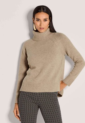 Vrouw in een beige coltrui en zwarte met patronen bedrukte broek, poseert met één hand op de heup tegen een effen achtergrond.