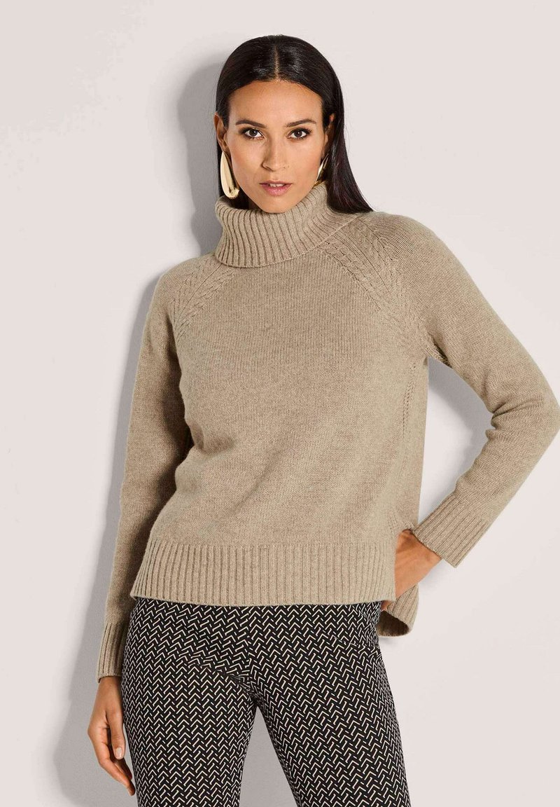 Frau in beigem Rollkragenpullover und schwarzen gemusterten Hosen, die mit einer Hand auf der Hüfte vor einem schlichten Hintergrund posiert.