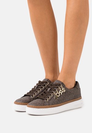 Zapatillas Michael Kors de color marrón con estampado y charms dorados en los cordones, con suelas gruesas blancas, usadas sobre pies descalzos contra un fondo blanco.