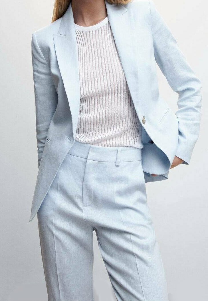 Personne portant un blazer bleu clair et un pantalon assorti avec un haut en maille côtelée blanche sur fond uni.